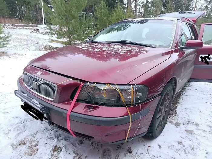 Red car with a broken headlight held by rubber bands and a strap as a creative if it's stupid but it works example in snow.