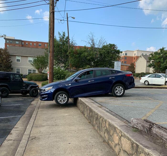 Blue sedan stuck halfway off a raised parking area, illustrating dumb drivers in a city setting.