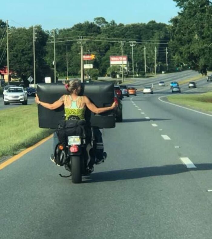 Person on motorcycle carrying large TV on busy road, illustrating dumb drivers shamed for unsafe behavior.
