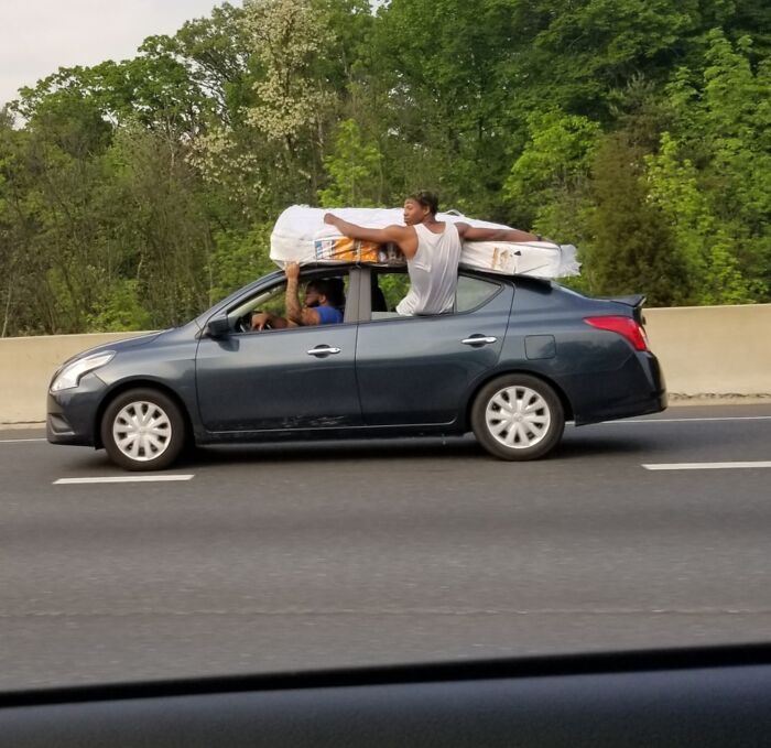 Man dangerously holding large mattress on car roof while driving, showcasing one of the dumb drivers on the road.
