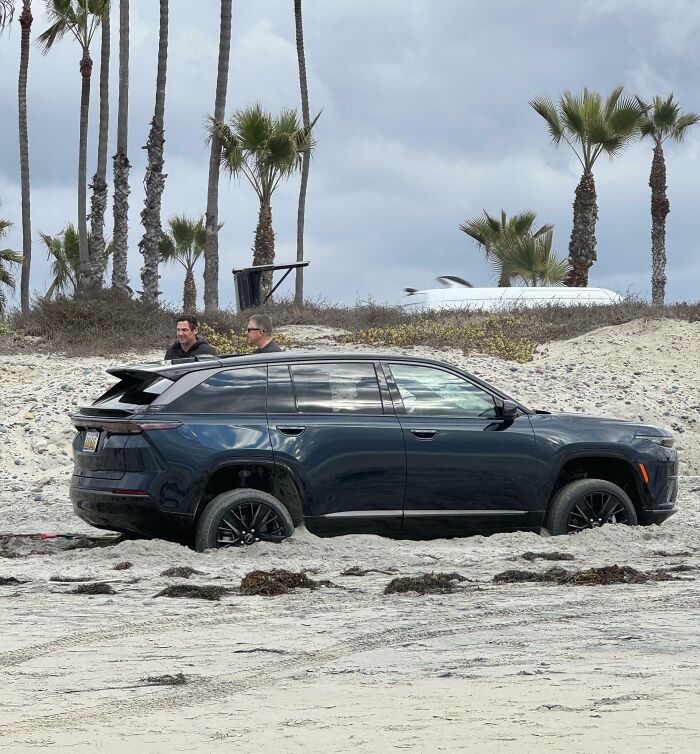 SUV stuck in deep sand on beach with two men nearby, illustrating dumb drivers caught in tricky situations.