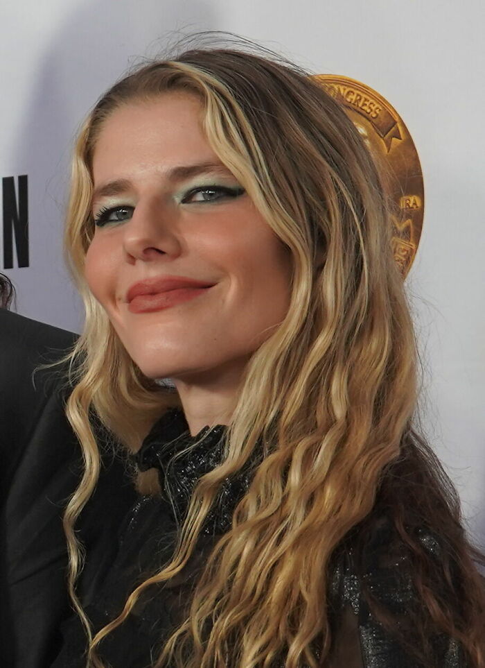 Katy Rose smiling at an event, with long wavy blonde hair and wearing a black outfit, showcasing her bio and career highlights.