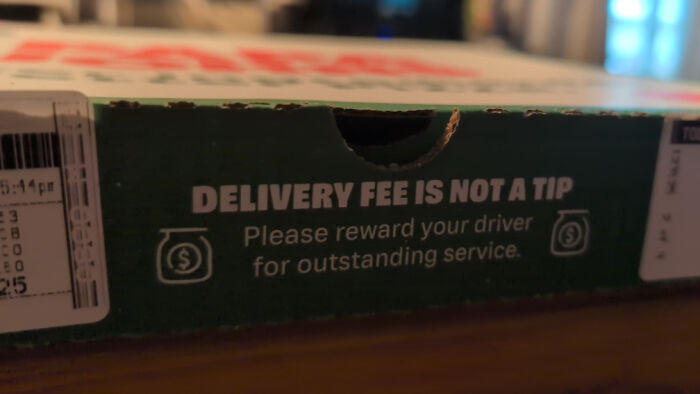Close-up of pizza box warning that delivery fee is not a tip, highlighting issues related to consumerism and service industry.