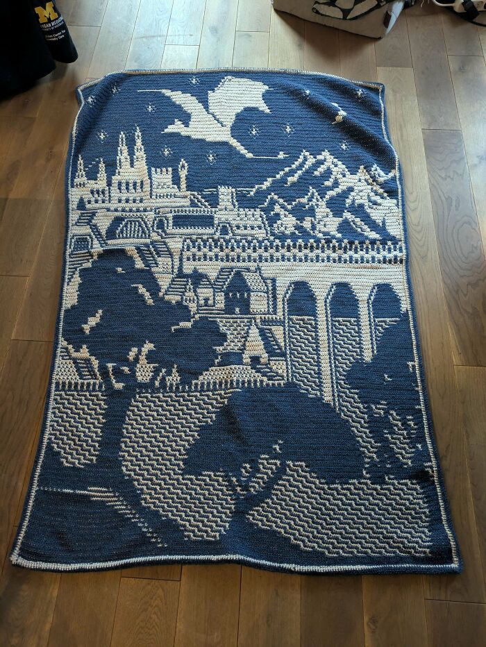 A stunning blue and white crocheted blanket featuring a fantasy landscape of castles, mountains, and a dragon. Awesome crocheted art.