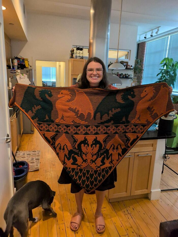 A smiling woman proudly displays her awesome crocheted shawl featuring a dragon pattern, a truly impressive crocheted item.