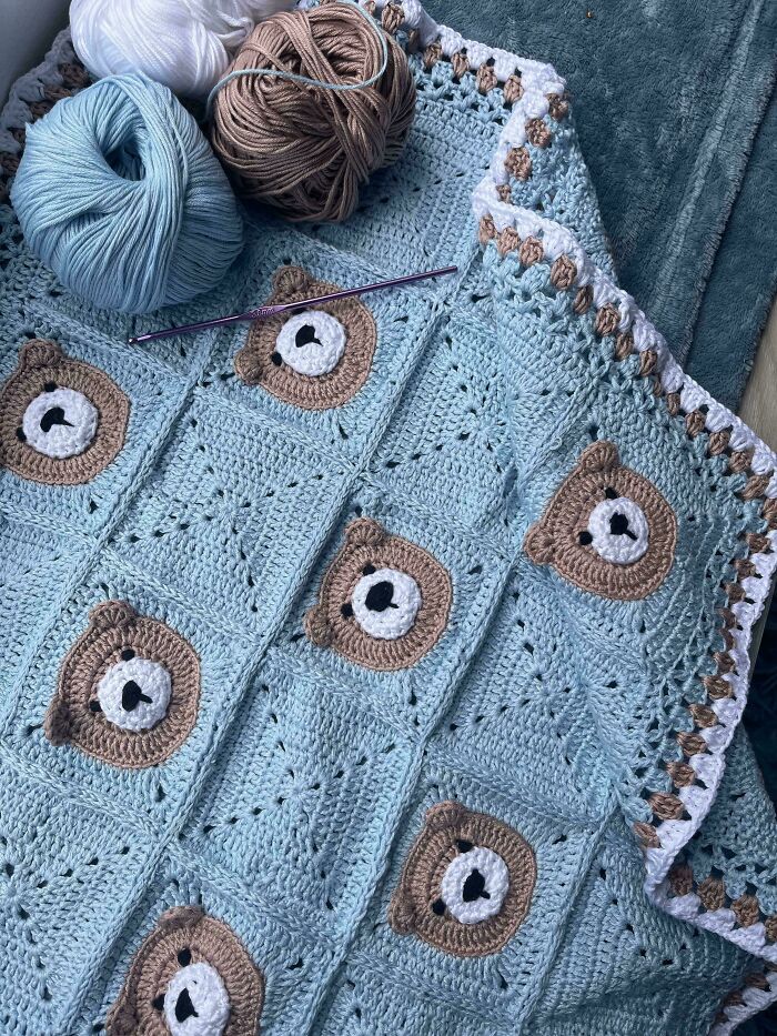 A light blue crocheted blanket with bear face squares, yarn balls, and a crochet hook. Awesome crocheted item.