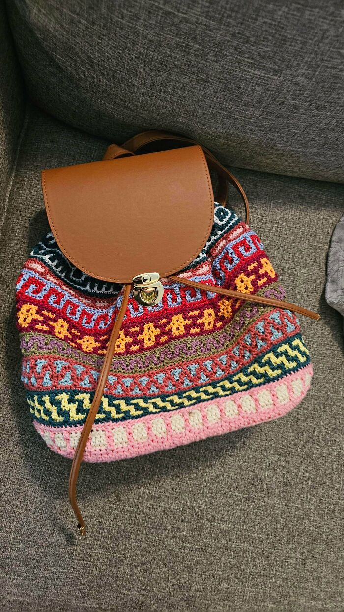 A beautifully crocheted backpack with a brown leather flap and intricate colorful patterns, an awesome crocheted item.