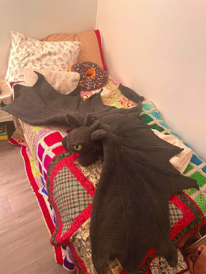 An awesome crocheted black dragon blanket with green eyes and large wings lays on a colorful patchwork quilt.