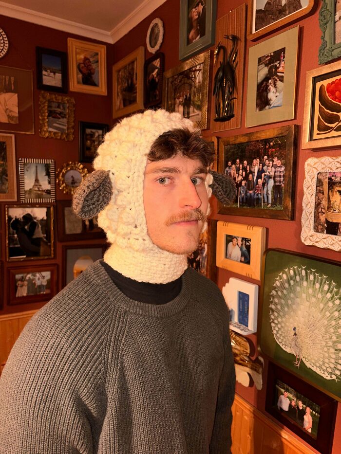 A man in a crocheted sheep balaclava, a truly awesome crocheted item, standing in a room with many framed pictures.