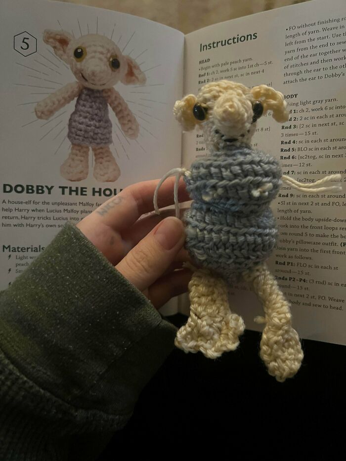 A hand holding a crocheted Dobby from Harry Potter, with the crochet pattern book open in the background. Awesome crocheted item.