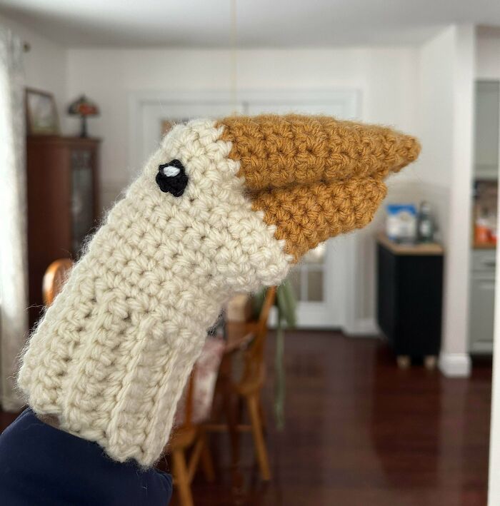 A hand wearing a crocheted bird puppet, white with a brown beak and black eye. This awesome crocheted item wowed the internet.