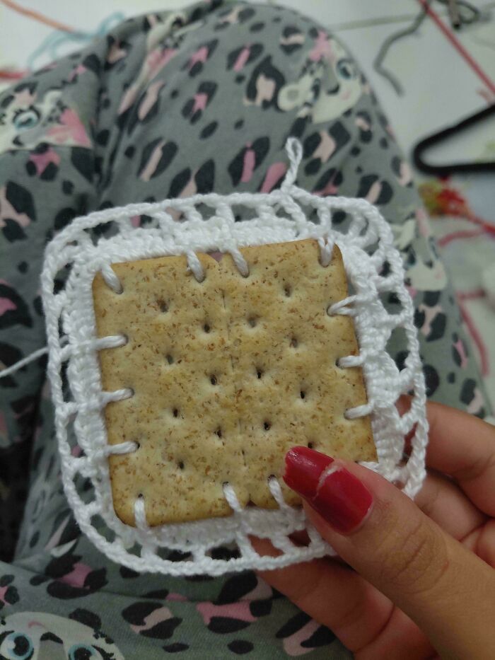 A hand with red nail polish holds a cracker stitched into a white crocheted square, a truly awesome crocheted item.