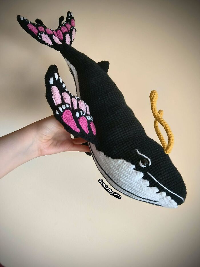A hand holding an awesome crocheted black whale with pink and white fins and tail, wowing the internet.