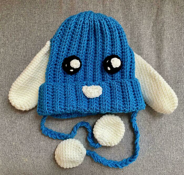 A blue crocheted beanie with white bunny ears, black eyes, and a small white mouth, showcasing awesome crocheted work.