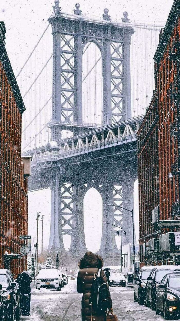 A large, eerie bridge stands tall between snow-covered buildings, with a person walking away in the foreground.