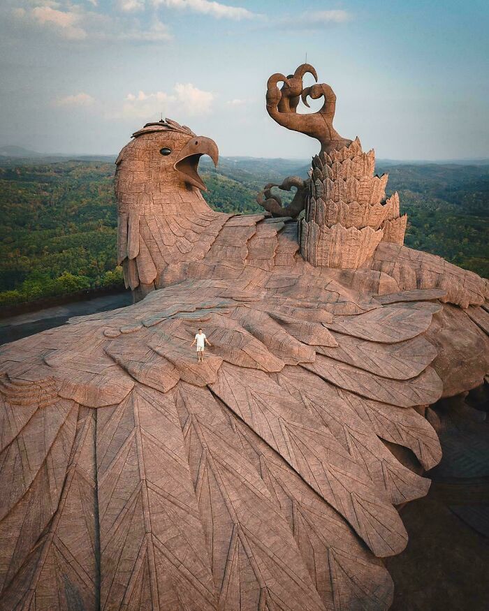 A person stands on the back of an extremely large, scary, and eerie eagle statue carved into a mountain.