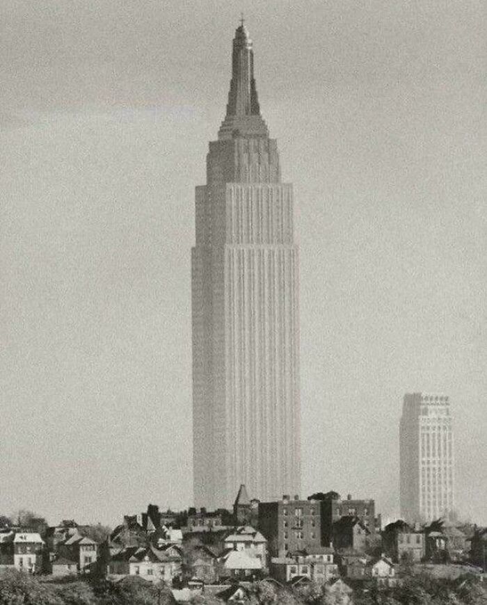 A sepia-toned image of an extremely large skyscraper towering over smaller buildings, creating a scary and eerie scale.