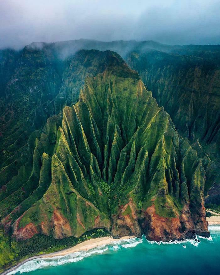 An aerial view of an extremely large, green, jagged mountain meeting the ocean, showing the scary and eerie scale.