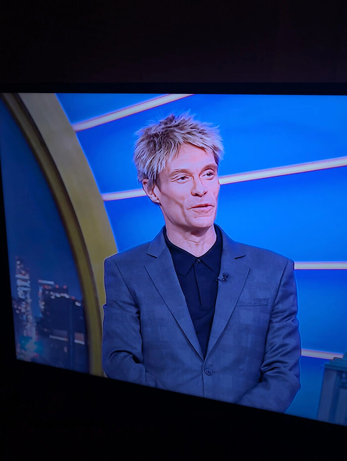 A man with spiky blonde hair, wearing a gray suit, exemplifies unfortunate haircuts captured from a TV screen.