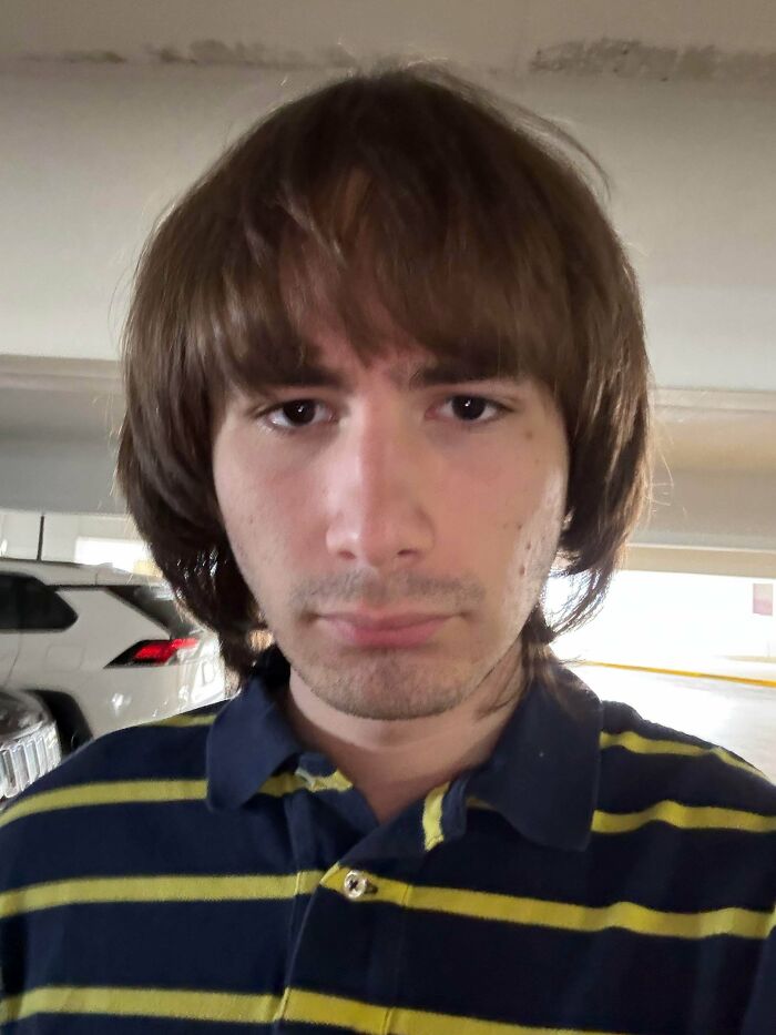 A young man with an unfortunate shaggy brown haircut and bangs, wearing a striped polo shirt.