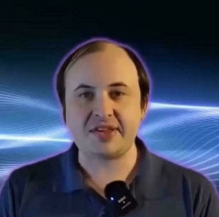 A man with an unfortunate haircut and a receding hairline smiles at the camera against a blue background.