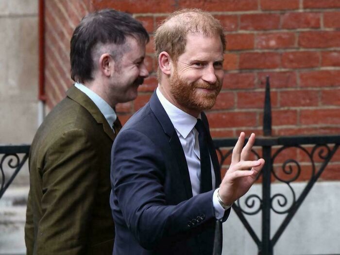 Prince Harry smiles at the camera, while a man with an unfortunate haircut stands behind him.