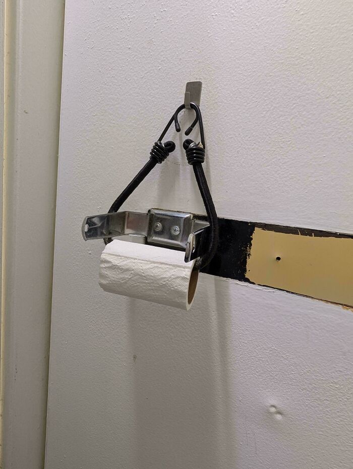 Improvised redneck engineering toilet paper holder made from metal bracket and bungee cords hanging on a wall hook.