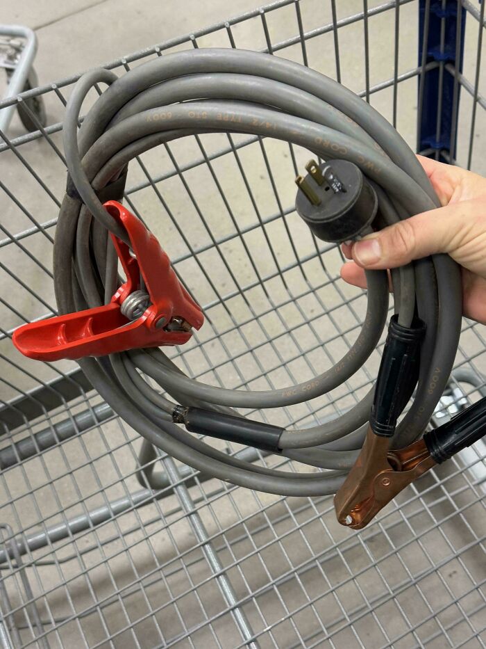 Hand holding a coiled jumper cable with a red clamp and a three-prong plug in a shopping cart, showing redneck engineering.