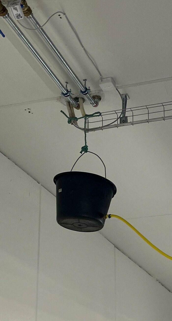 Improvised redneck engineering bucket hanging from ceiling pipe with yellow hose attached inside an industrial setting.