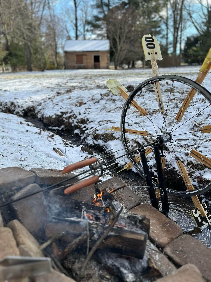 Improvised redneck engineering water wheel made from a bicycle wheel and wooden paddles beside a fire pit in a snowy yard.
