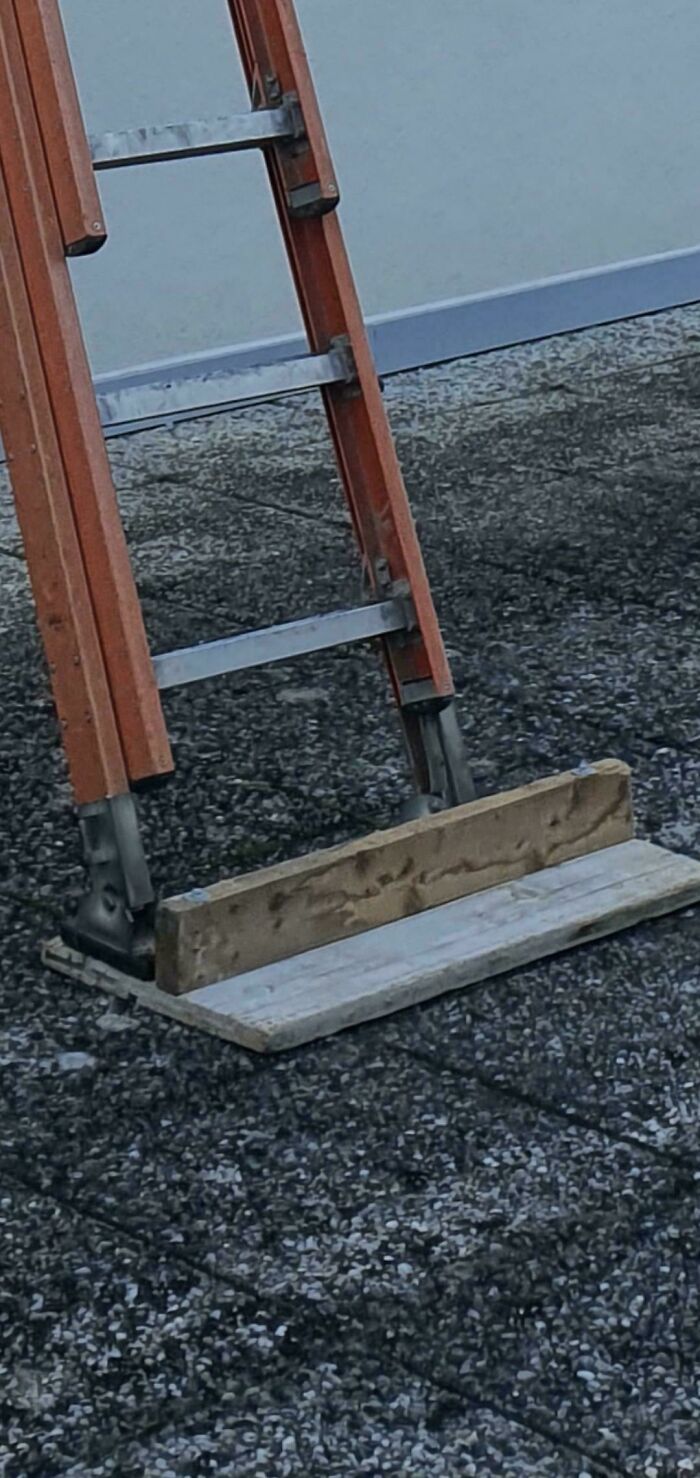 Wooden ladder with a makeshift base made of planks balancing on uneven gravel surface redneck engineering example.