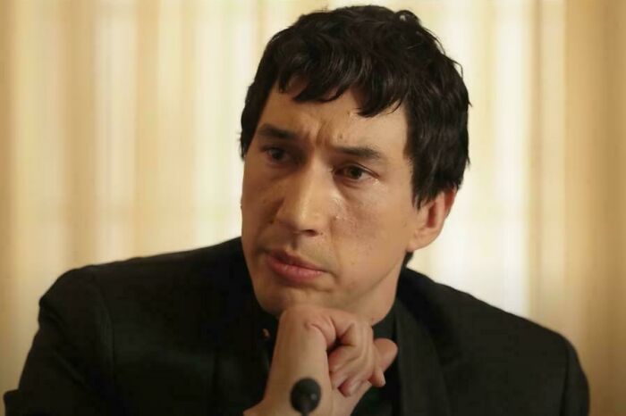 Man with dark hair and black jacket thoughtfully resting chin on hand in a scene from absurd movie and TV details.