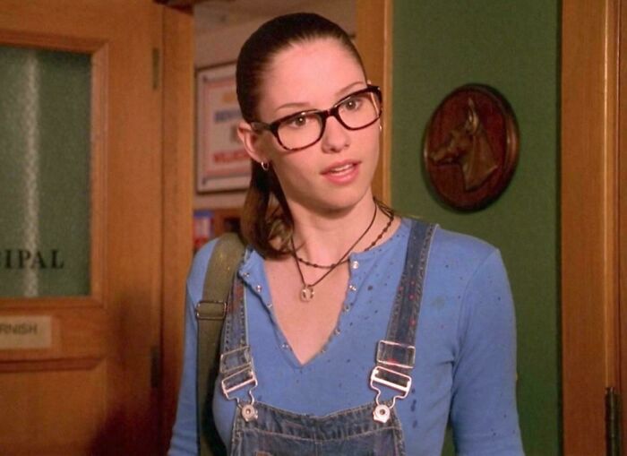 Young woman in glasses and denim overalls standing indoors, representing hilariously absurd movie and TV details.