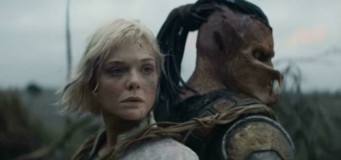 Woman with short blonde hair and creature with mask looking in opposite directions in a movie scene showing absurd TV details.