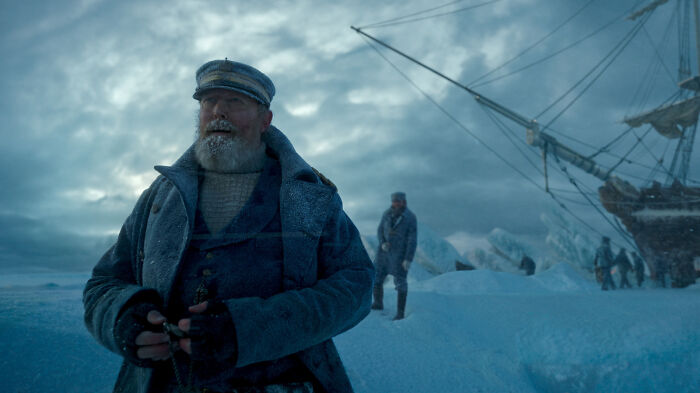 Man dressed as a ship captain standing in icy landscape with a large ship in the background, absurd movie detail scene