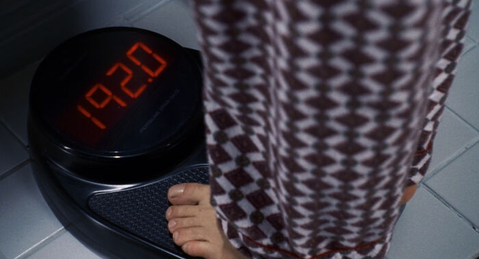 Person in patterned pants standing on digital scale showing 192.0 in a scene depicting absurd movie and TV details.