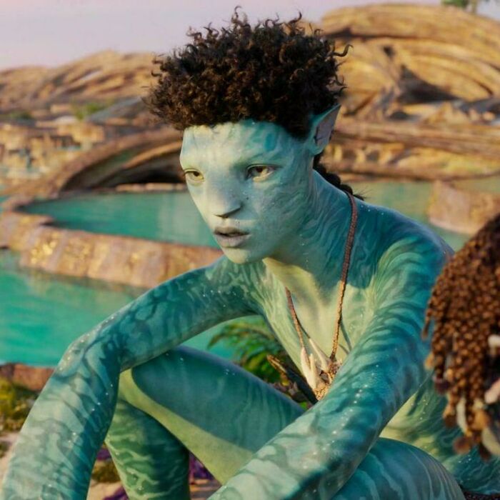 Blue-skinned humanoid character with curly hair sitting near water in a scene featuring absurd movie and TV details.