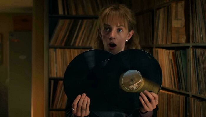 A woman holding two overlapping vinyl records in front of shelves filled with records, showing a surprised expression.
