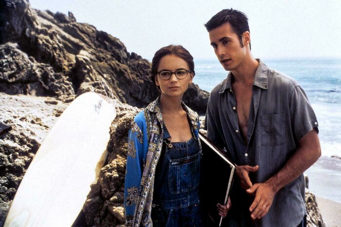 Two young adults on a rocky beach, reflecting on ’90s childhood memories with surfboard and casual clothing.