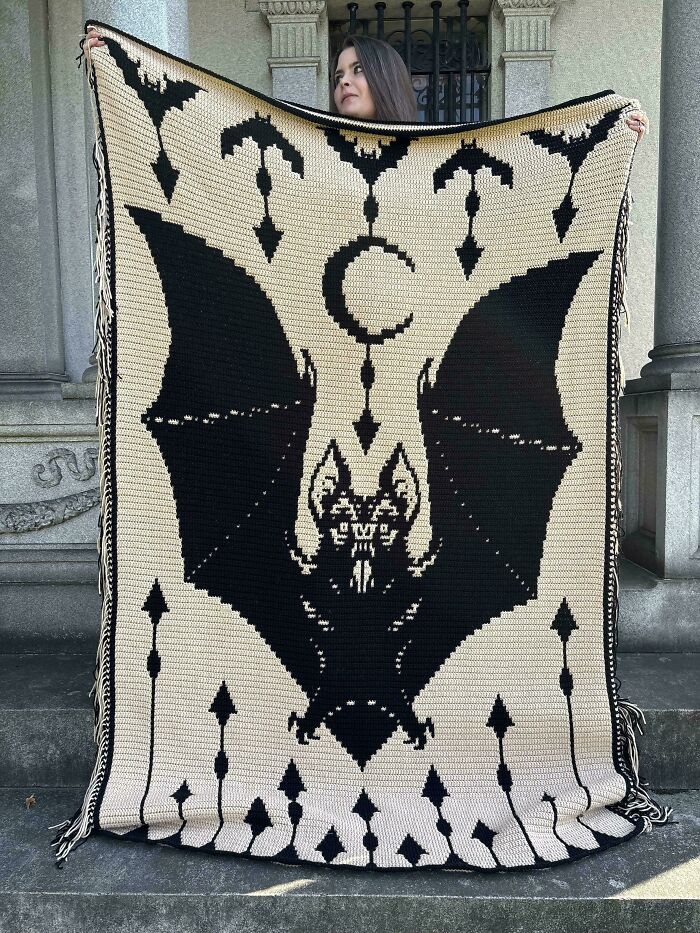 Woman holding a large crocheted blanket with a black bat design, showcasing a DIY craft win with detailed patterns.