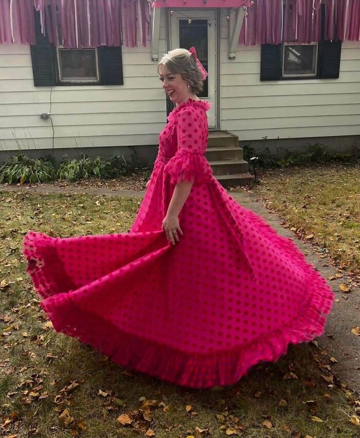 Woman wearing a bright pink polka dot dress spinning outside a house, showcasing her DIY wins with a joyful expression.