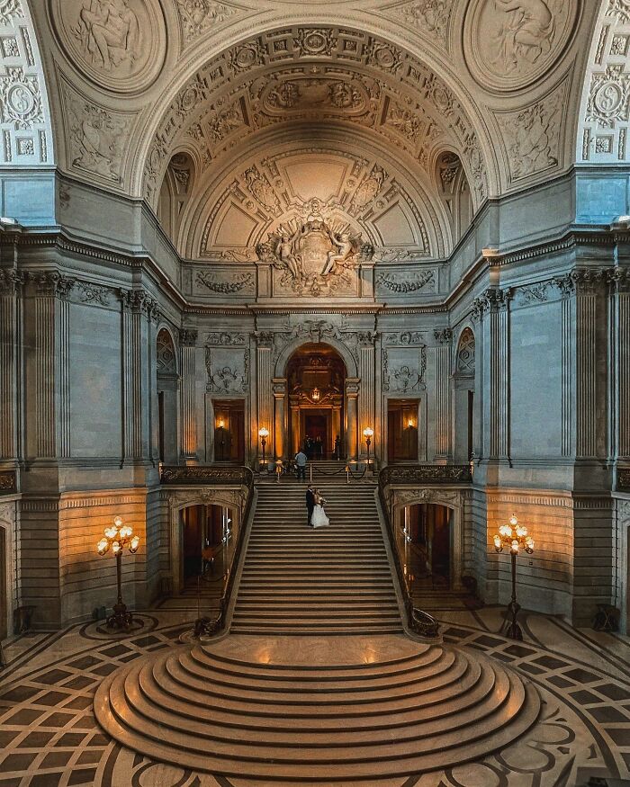 A grand hall with beautiful architecture, a sweeping staircase, and a couple standing on the landing.
