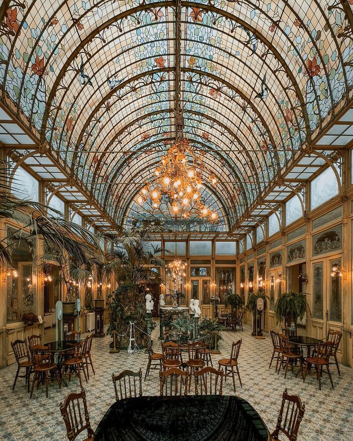 A vast room with an arched stained-glass ceiling, lush plants, and many tables and chairs, showcasing beautiful architecture.