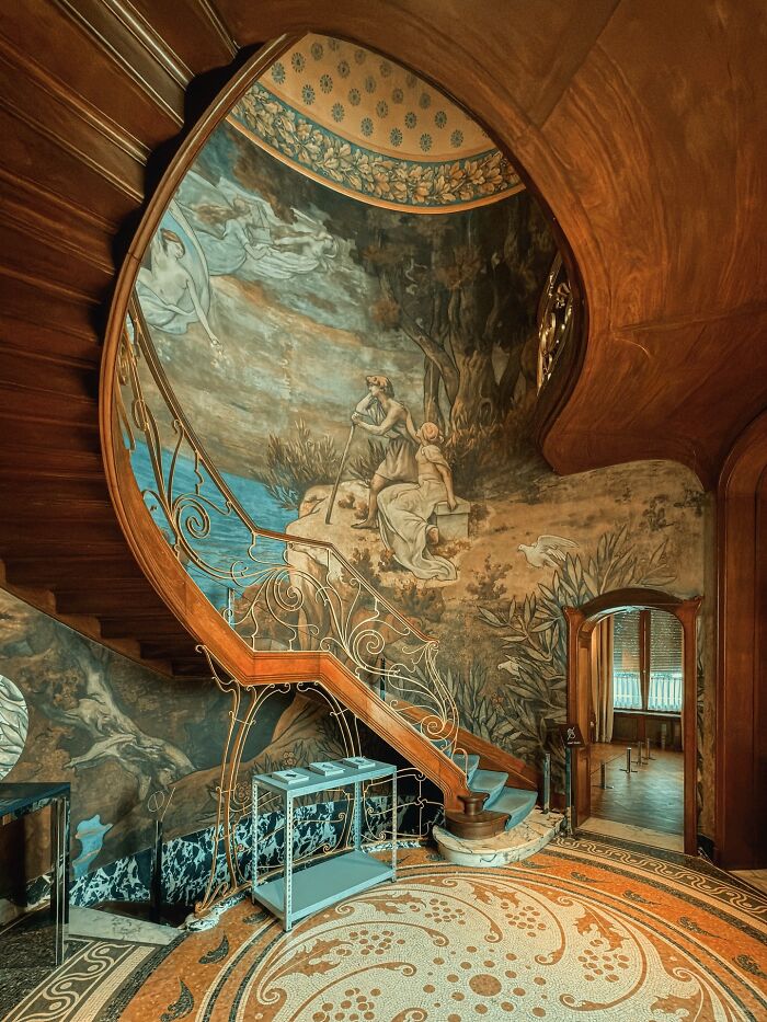 A grand staircase with a detailed mural along the wall and decorative railings, an example of beautiful architecture.