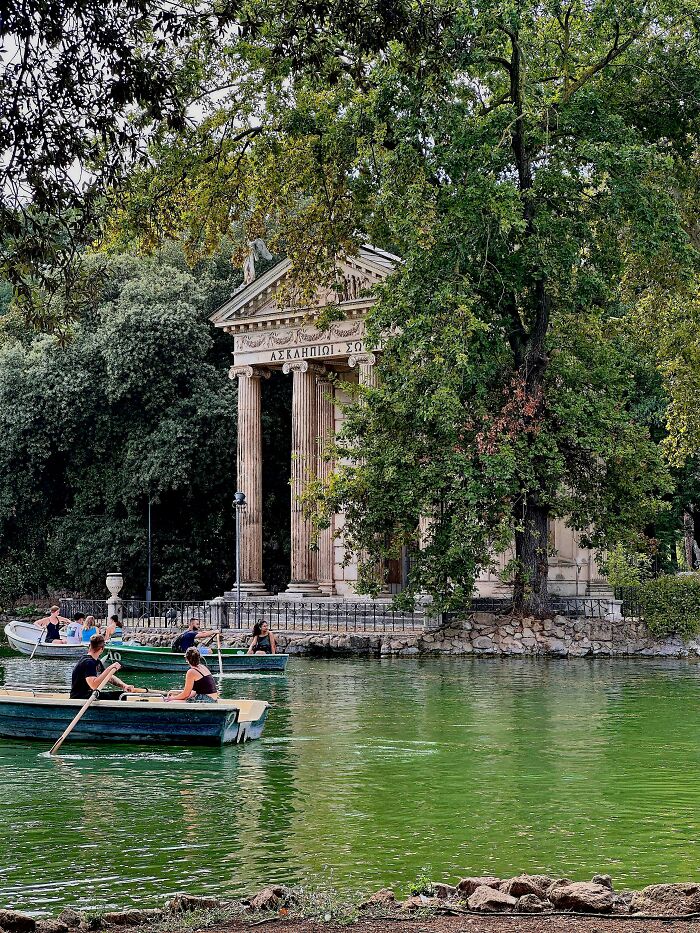 A beautiful classical architecture building with columns overlooking a tranquil green lake with people rowing boats, showcasing beautiful architecture.