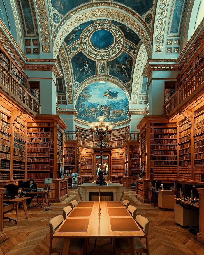 Grand library with towering bookshelves, intricate ceiling art, and a long central table showcasing beautiful architecture.