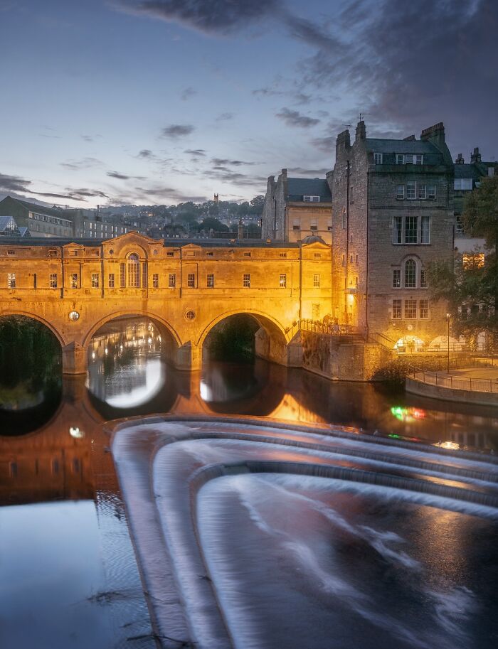 An illuminated stone bridge and buildings reflect on water at dusk, exemplifying beautiful architecture.