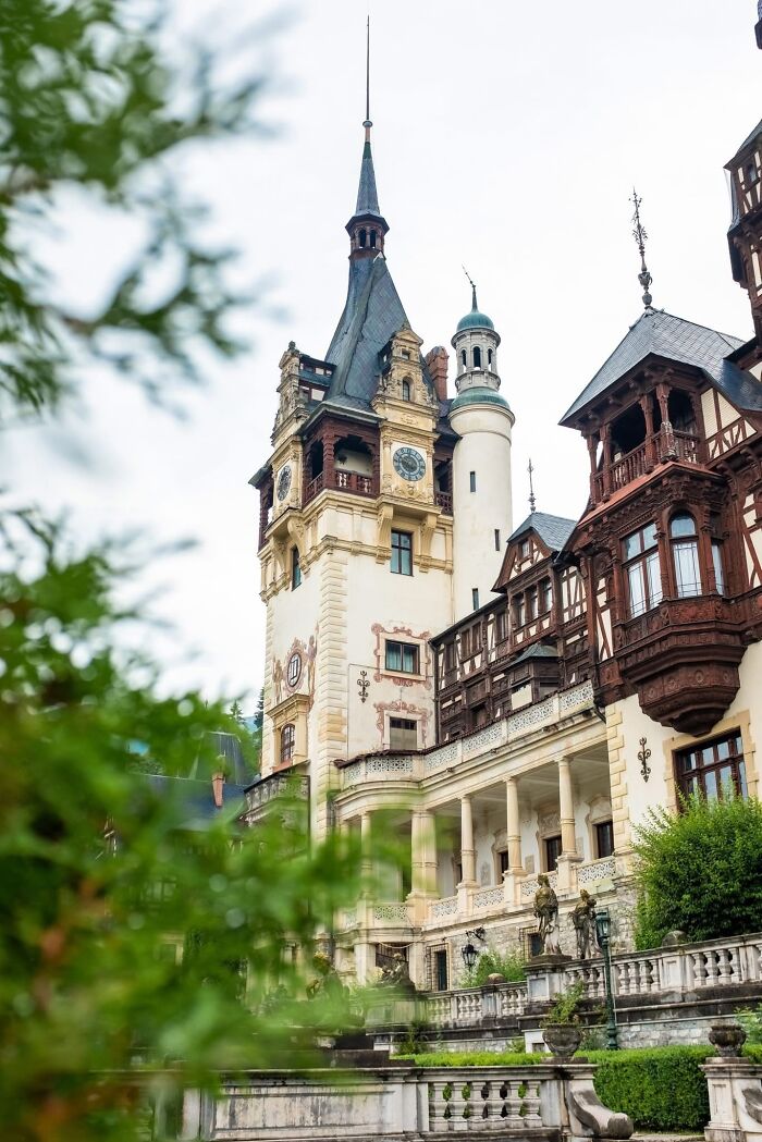 Intricate architecture of a historical castle with towers and ornate balconies, seen through green foliage.