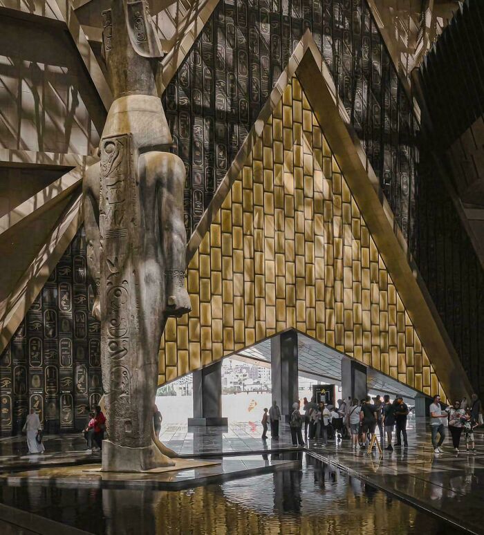 Grand statue in a modern museum with striking geometric architecture and ancient hieroglyphics.