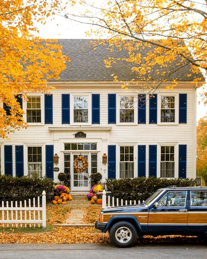 Charming white colonial architecture with blue shutters and vibrant fall decor, with a vintage car.